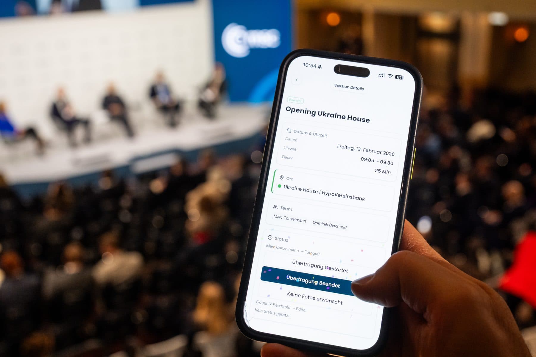 A phone held up in the conference hall: the session detail view in VAULO Events with date, place, team and status — the panel on stage blurred behind