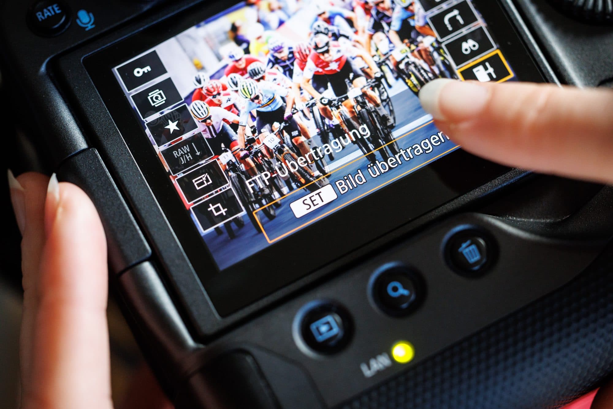 Camera display at a cycling race: the "FTP transfer — transfer image" menu sits over the photo of the peloton, a finger taps transfer