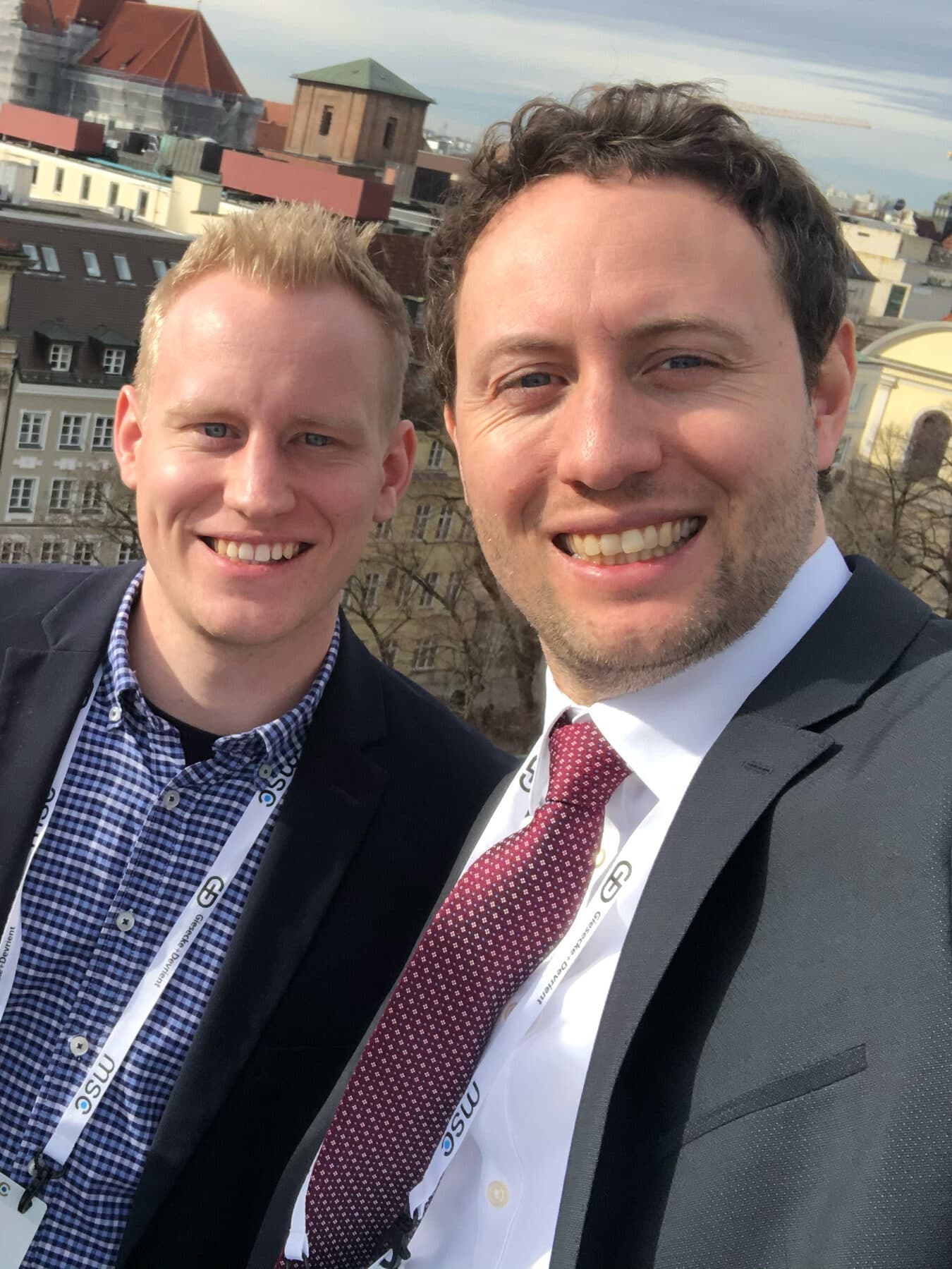 Sebastian Küsters and Marc Conzelmann, a selfie with conference lanyards in front of the roofs and church towers of Munich's old town