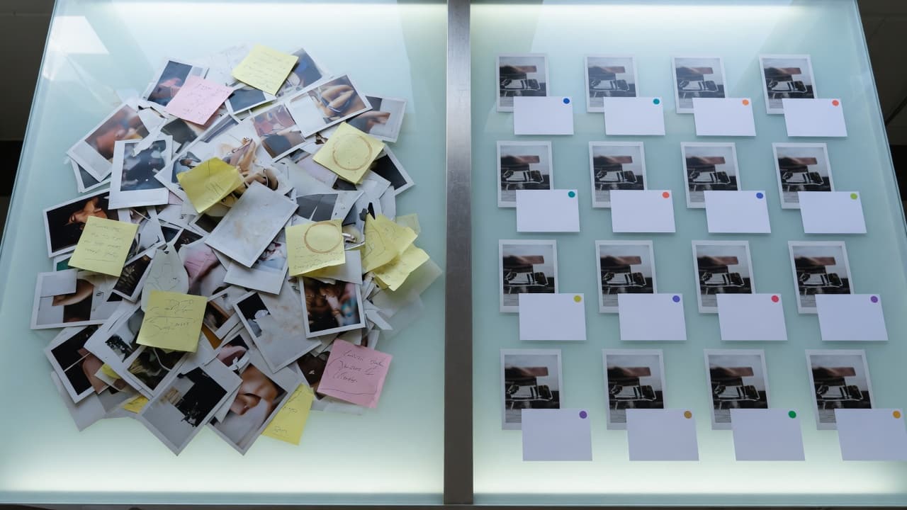 AI-generated still life: on the left an unsorted stack of photo prints with sticky notes, on the right the same prints arranged in a grid, each with an index card and colored marker dots